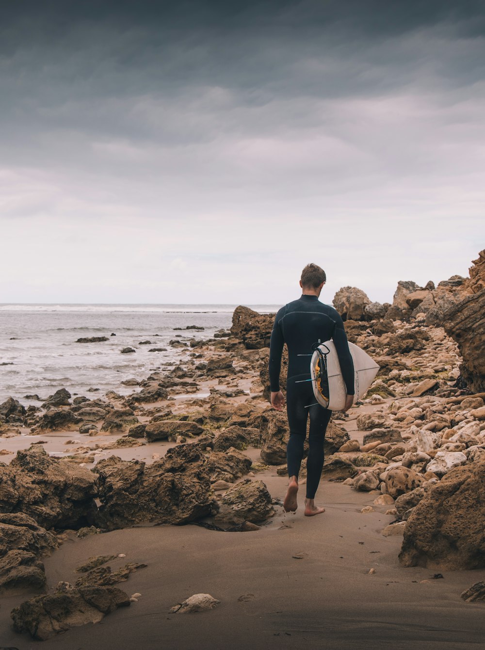Un hombre con traje de neopreno cargando una tabla de surf en una playa rocosa