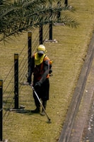 A person is using a grass trimmer on a well-maintained lawn next to a path. There are black metal posts with yellow markers connected by cables running alongside the lawn. The person is wearing protective clothing including a hat, gloves, and a face covering.