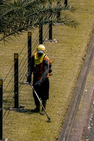 A person is using a grass trimmer on a well-maintained lawn next to a path. There are black metal posts with yellow markers connected by cables running alongside the lawn. The person is wearing protective clothing including a hat, gloves, and a face covering.