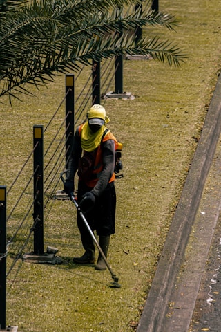 A person is using a grass trimmer on a well-maintained lawn next to a path. There are black metal posts with yellow markers connected by cables running alongside the lawn. The person is wearing protective clothing including a hat, gloves, and a face covering.