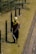 A person is using a grass trimmer on a well-maintained lawn next to a path. There are black metal posts with yellow markers connected by cables running alongside the lawn. The person is wearing protective clothing including a hat, gloves, and a face covering.