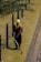 A person is using a grass trimmer on a well-maintained lawn next to a path. There are black metal posts with yellow markers connected by cables running alongside the lawn. The person is wearing protective clothing including a hat, gloves, and a face covering.