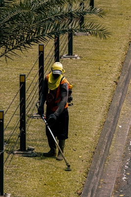 A person is using a grass trimmer on a well-maintained lawn next to a path. There are black metal posts with yellow markers connected by cables running alongside the lawn. The person is wearing protective clothing including a hat, gloves, and a face covering.