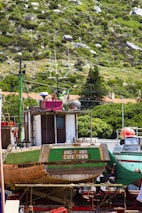 A large wooden fishing boat named 'Ang-Jerry Cape Town' is docked on a red stand, surrounded by a lush, green hillside. The boat shows signs of wear, with visible patches of paint and wood grain. In the background, another boat with a red and white color scheme can be seen, and greenery from nearby trees frames the scene.