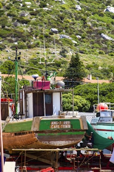 A large wooden fishing boat named 'Ang-Jerry Cape Town' is docked on a red stand, surrounded by a lush, green hillside. The boat shows signs of wear, with visible patches of paint and wood grain. In the background, another boat with a red and white color scheme can be seen, and greenery from nearby trees frames the scene.