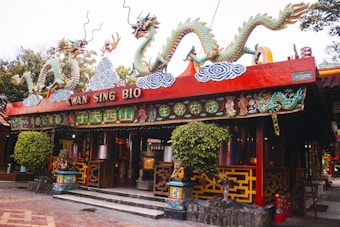 An ornate, colorful temple entrance adorned with intricate carvings and two large dragon sculptures on the roof. The building has traditional Asian architecture with bright red and green hues, intricate patterns, and Chinese characters. Lush greenery and decorative elements surround the entrance.