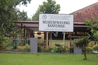 A building with a large sign that reads 'Museum Wayang Banyumas' stands behind a decorative fence. The museum is surrounded by greenery, including bushes and trees, under a clear sky. The architecture features a red tiled roof and a covered walkway leading to the entrance.
