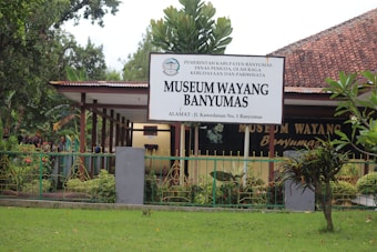 A building with a large sign that reads 'Museum Wayang Banyumas' stands behind a decorative fence. The museum is surrounded by greenery, including bushes and trees, under a clear sky. The architecture features a red tiled roof and a covered walkway leading to the entrance.