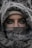 A person with expressive eyes is partially concealed behind a patterned fabric that covers most of the face and head. The intricate designs on the fabric are prominent, and the eyes convey a sense of mystery.