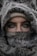 A person with expressive eyes is partially concealed behind a patterned fabric that covers most of the face and head. The intricate designs on the fabric are prominent, and the eyes convey a sense of mystery.