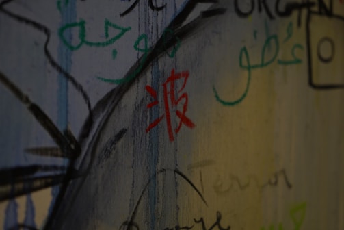 Various scripts and symbols are drawn on a wall with a mix of colors like red, green, and black. The writing includes different styles, possibly from different languages, creating an artistic and graffiti-like effect.