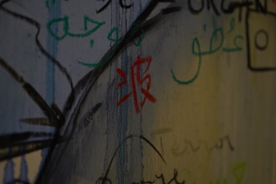 Various scripts and symbols are drawn on a wall with a mix of colors like red, green, and black. The writing includes different styles, possibly from different languages, creating an artistic and graffiti-like effect.