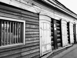 A rustic wooden building with several closed shutters and visible wooden texture. The structure has a weathered appearance, with vertical and horizontal wooden planks and an old window with bars.