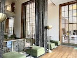 A modern interior space featuring large windows with geometric patterns and marble columns. The room has green upholstered benches and contemporary metallic floor lamps. A wooden floor complements the light neutral walls, creating a spacious and airy atmosphere.