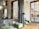 A modern interior space featuring large windows with geometric patterns and marble columns. The room has green upholstered benches and contemporary metallic floor lamps. A wooden floor complements the light neutral walls, creating a spacious and airy atmosphere.