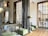 A modern interior space featuring large windows with geometric patterns and marble columns. The room has green upholstered benches and contemporary metallic floor lamps. A wooden floor complements the light neutral walls, creating a spacious and airy atmosphere.