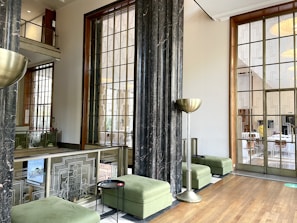A modern interior space featuring large windows with geometric patterns and marble columns. The room has green upholstered benches and contemporary metallic floor lamps. A wooden floor complements the light neutral walls, creating a spacious and airy atmosphere.