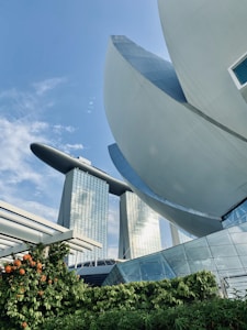 Futuristic architecture is captured with high-rise buildings featuring a unique design that resembles a ship balanced on top of three towers. In the foreground, a structure with curved, petal-like elements is visible alongside lush greenery and vibrant orange flowers. The sky is clear with minimal clouds, providing a backdrop for the modern structures.
