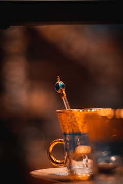 A close-up of an ornate glass cup featuring a delicate stick or spoon with a decorative end, resting on an elegant saucer. The background is softly blurred, emphasizing the cup's intricate details.