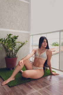 A woman is sitting on a green mat in a balcony area. She is wearing a bikini with crocheted edging. Beside her, there is a potted plant with green leaves. The balcony has a tiled floor and a view of rooftops and trees in the background.