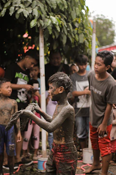 Children and adults making mud crafts together under a shaded tree.