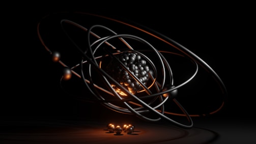 A dark, abstract, and futuristic representation of an atom, featuring a cluster of metallic spheres at the center surrounded by orbiting rings. The orange and black lighting adds a dramatic effect, emphasizing the spherical forms and giving a sense of depth.