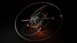 A 3D model of an atom with metallic and glossy elliptical rings surrounding a cluster of black and reflective spheres at the center, set against a dark background. Warm orange highlights accentuate the structure and add contrast to the composition.