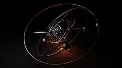 A 3D model of an atom with metallic and glossy elliptical rings surrounding a cluster of black and reflective spheres at the center, set against a dark background. Warm orange highlights accentuate the structure and add contrast to the composition.