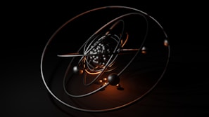 A 3D model of an atom with metallic and glossy elliptical rings surrounding a cluster of black and reflective spheres at the center, set against a dark background. Warm orange highlights accentuate the structure and add contrast to the composition.