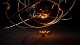 A 3D representation of an atomic structure with orbiting electrons. The nucleus is composed of tightly packed spheres and surrounded by several rings with individual spheres representing electrons. A warm glow illuminates the nucleus, casting shadows and reflections.