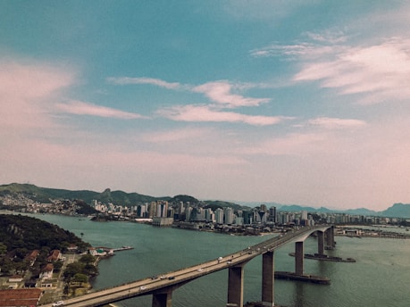 A large bridge extends across a wide body of water, connecting two land masses. To the left, residential areas with houses and greenery are visible, while to the right, a cityscape with numerous high-rise buildings is seen in the background. Rolling hills and mountains can be seen in the distance under a partly cloudy sky.