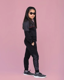 A young girl stands confidently against a solid pink background. She is wearing a dark tracksuit with subtle details, black sunglasses, and sports shoes. Her hair is styled neatly, and she has a calm, composed expression.