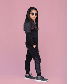A young girl stands confidently against a solid pink background. She is wearing a dark tracksuit with subtle details, black sunglasses, and sports shoes. Her hair is styled neatly, and she has a calm, composed expression.