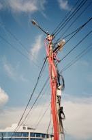 A utility pole is cluttered with numerous black and red cables and wires in various directions, set against a clear blue sky. Part of a modern building with reflective windows is visible in the background.