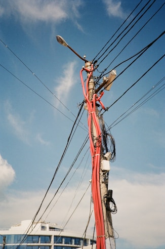 A utility pole is cluttered with numerous black and red cables and wires in various directions, set against a clear blue sky. Part of a modern building with reflective windows is visible in the background.