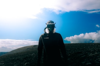 A person wearing a protective headset and mask stands on a rocky terrain under a bright blue sky with scattered clouds.