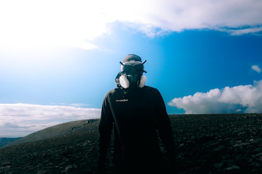A person wearing a protective headset and mask stands on a rocky terrain under a bright blue sky with scattered clouds.