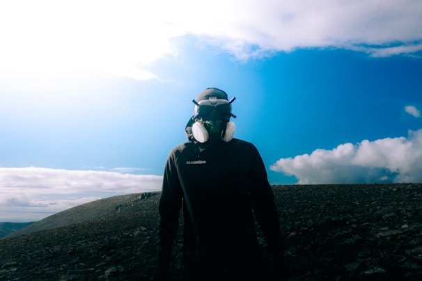 A person wearing a protective headset and mask stands on a rocky terrain under a bright blue sky with scattered clouds.