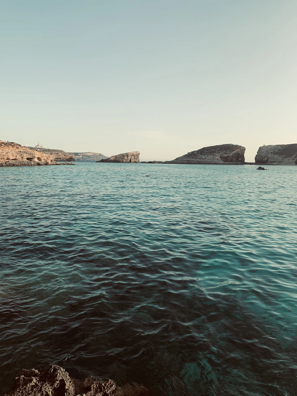 The image depicts a serene coastal scene with a deep blue ocean stretching out to meet a rocky shore on the horizon. The sky above is a light blue, providing a beautiful backdrop for the tranquil waters below. In the foreground, there are several small rocks scattered across the water's surface, adding texture and depth to the landscape. A few boats can be seen in the distance, further enhancing the sense of tranquility and solitude that this picturesque setting offers.