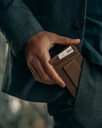 A person dressed in a tailored, dark-colored suit is placing a leather wallet with visible currency, specifically a twenty-dollar bill, into their pocket. The focus is on the hand and wallet, conveying a sense of sophistication and care.