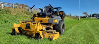 A yellow industrial lawn mower sits on a freshly cut grassy field. It's positioned prominently in the foreground, with large black tires and a robust metal frame. In the background, a green hillside and a clear blue sky are visible. To the right, a pole and signage for a lawn equipment store can be seen in the distance.