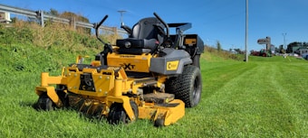 A yellow industrial lawn mower sits on a freshly cut grassy field. It's positioned prominently in the foreground, with large black tires and a robust metal frame. In the background, a green hillside and a clear blue sky are visible. To the right, a pole and signage for a lawn equipment store can be seen in the distance.
