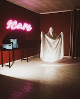 A room with dim lighting features a corner with a ghostly figure draped in a white sheet, standing beside a table. Above the table, neon pink letters spell out 'Scare.' The walls are a dark red, adding to the eerie atmosphere. Nearby, two blue boxes rest on the table, casting shadows.
