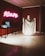 A room with dim lighting features a corner with a ghostly figure draped in a white sheet, standing beside a table. Above the table, neon pink letters spell out 'Scare.' The walls are a dark red, adding to the eerie atmosphere. Nearby, two blue boxes rest on the table, casting shadows.