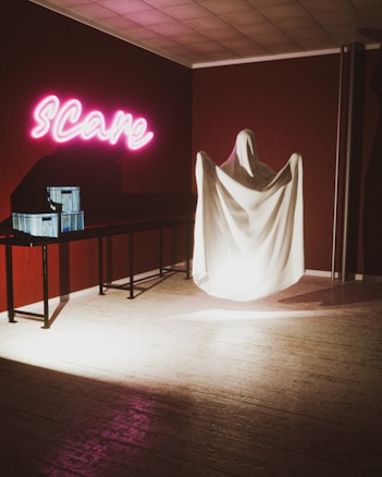 A room with dim lighting features a corner with a ghostly figure draped in a white sheet, standing beside a table. Above the table, neon pink letters spell out 'Scare.' The walls are a dark red, adding to the eerie atmosphere. Nearby, two blue boxes rest on the table, casting shadows.