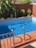 A small rectangular swimming pool with clear blue water is surrounded by terracotta tiles. There is a metal handrail for steps leading into the pool. Green leafy plants are against a tall, muted red wall in the background, providing a bit of lush contrast.