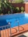 A small rectangular swimming pool with clear blue water is surrounded by terracotta tiles. There is a metal handrail for steps leading into the pool. Green leafy plants are against a tall, muted red wall in the background, providing a bit of lush contrast.