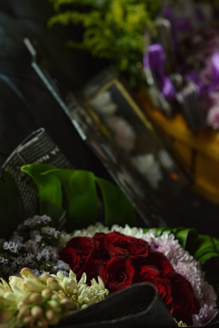 A bouquet comprising vibrant red roses, pale pink lilies, and small purple flowers, complemented by lush green leaves. The composition is artfully arranged against a dark background, creating a striking contrast and highlighting the rich colors of the flowers.