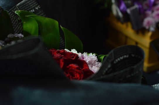 A bouquet wrapped in dark, textured paper with fresh green leaves, vibrant red roses, and delicate pink flowers is arranged in an inviting manner. In the background, a wooden crate holds other floral arrangements with hints of purple. The overall setting is dimly lit, adding a sense of mystery and focus to the flowers.