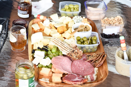 A charcuterie board with various types of cheeses, sliced meats such as salami and prosciutto, green and black olives, crackers, and a variety of spreads is laid out on a table. Accompanying containers hold nuts, dried fruits, dips, and pickles. Jars of preserves and an assortment of condiments are placed nearby, creating an inviting and colorful spread for a gathering or meal.